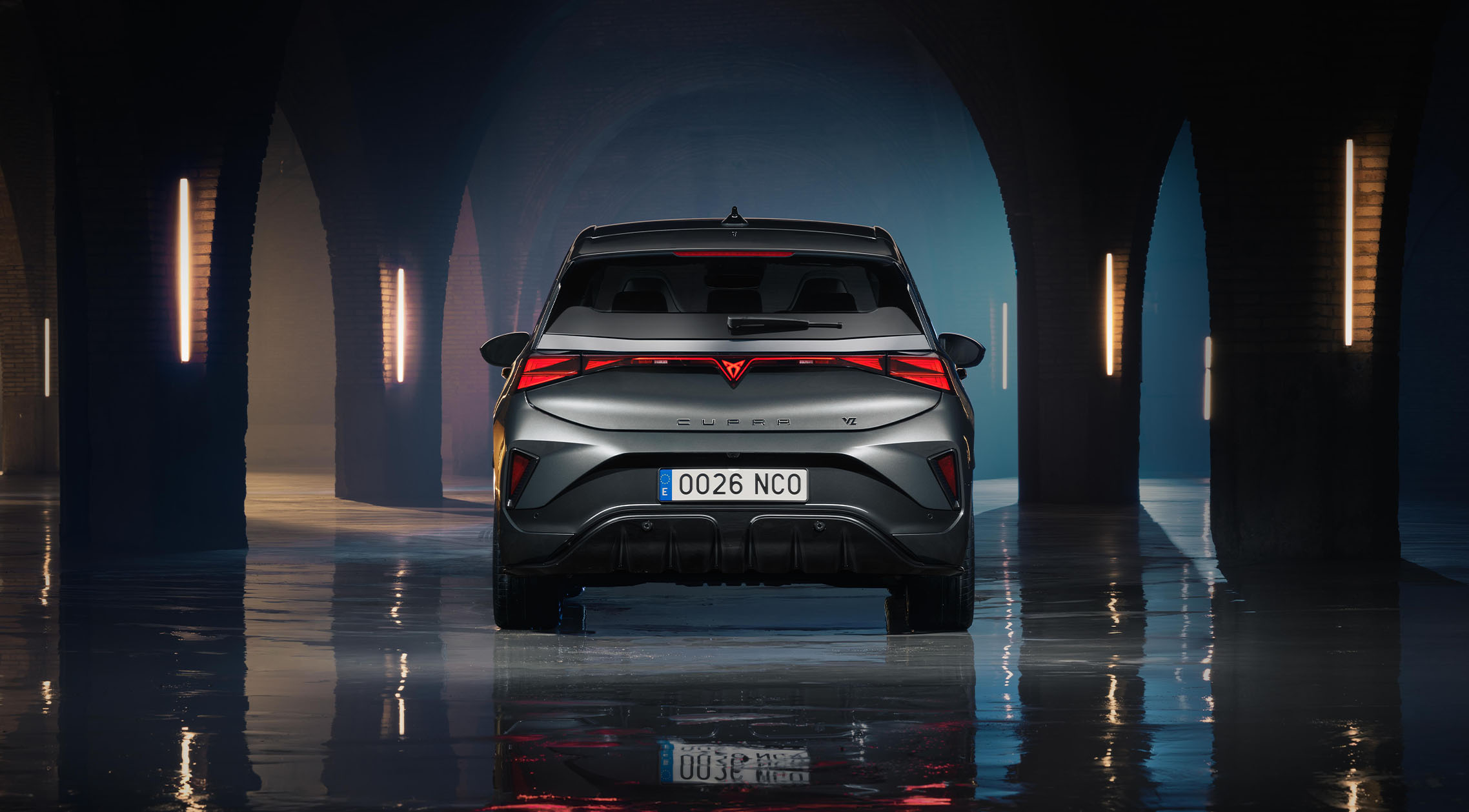 Vue arrière directe d’une CUPRA Born VZ en gris Timanfaya, stationnée dans un espace industriel sombre et voûté, avec des arches en pierre et un éclairage néon vertical. L’image met en valeur le bandeau lumineux arrière à LED continu, le logo CUPRA emblématique ainsi que le reflet du véhicule sur un sol en béton poli et mouillé.
