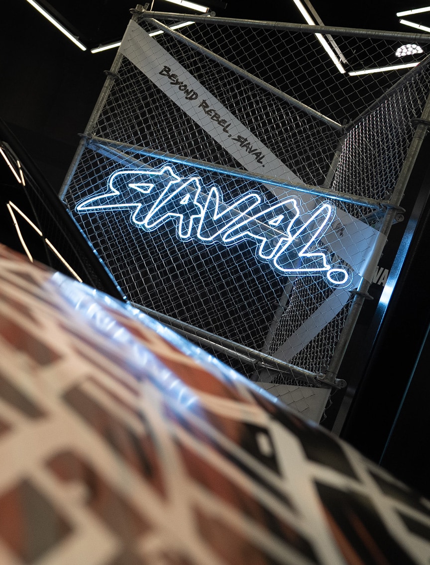 Une enseigne au néon affichant “RAVAL !” est installée sur un panneau de grillage, qui comporte également le texte “BEYOND REBEL, RAVAL.”. L’ensemble se trouve dans une pièce sombre et stylisée, avec des motifs lumineux géométriques.