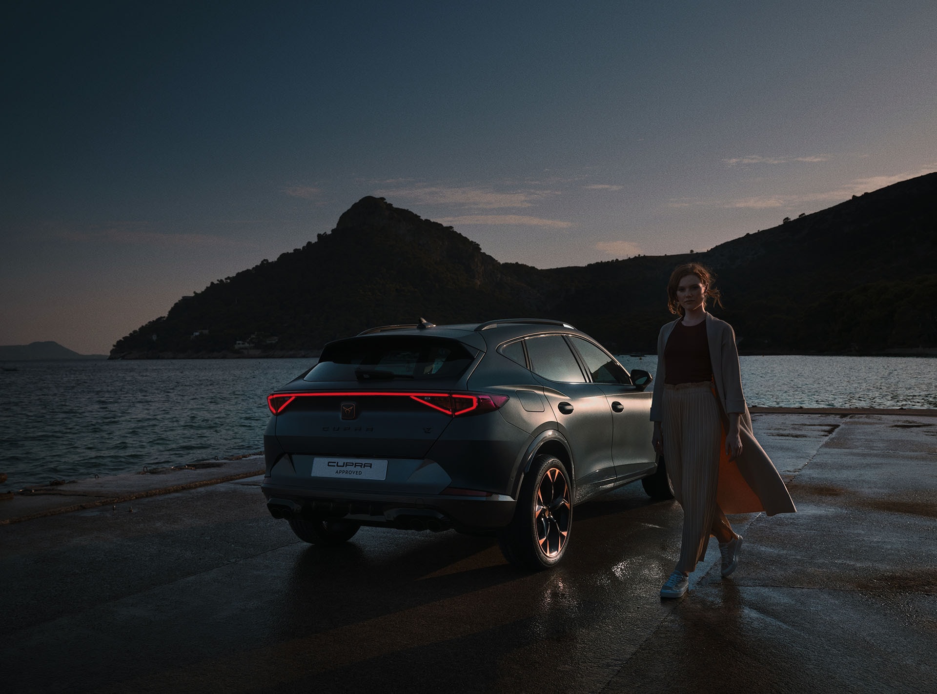Une CUPRA est garée sur un quai au bord de l'eau et une femme se tient à côté de la voiture et regarde le spectateur dans les yeux.