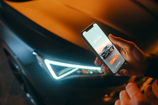 En arrière-plan, on voit un phare d'une CUPRA. Au premier plan, une main tient un téléphone portable sur l'écran duquel on peut voir le login de l'application CUPRA Connect.