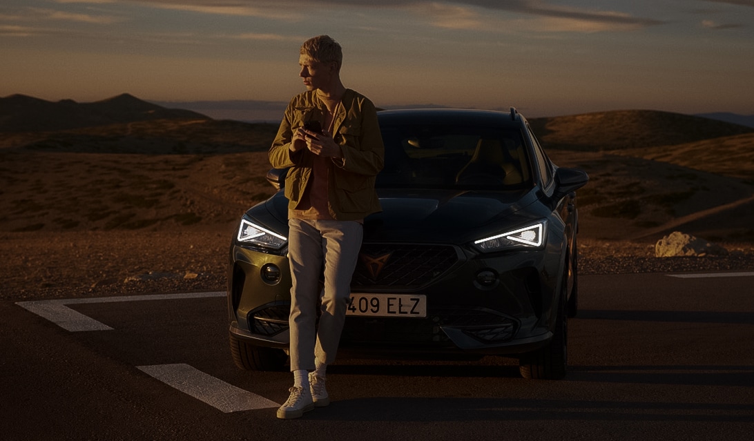 Un homme est appuyé à l'avant sur le capot d'une CUPRA et regarde au loin vers la droite.