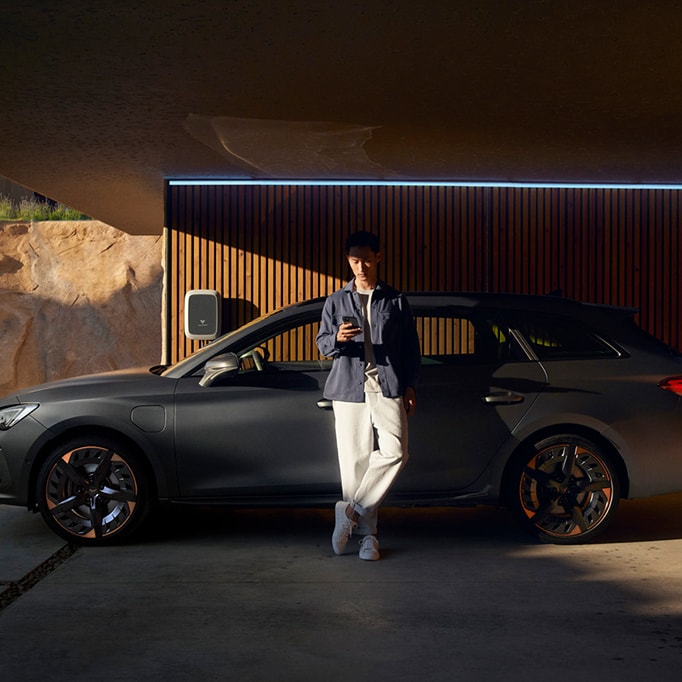 Une CUPRA se trouve sur le côté d'un mur de maison et est raccordée à une Wallbox. Un homme est appuyé contre la portière du conducteur et regarde son téléphone portable.
