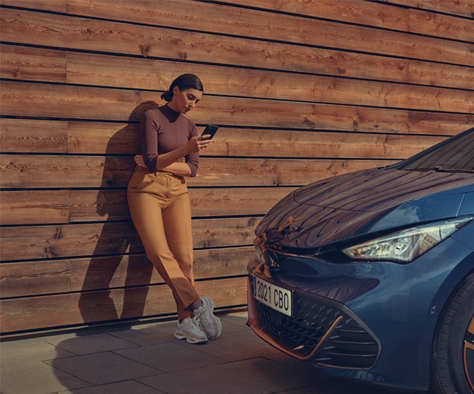 Femme adossée à un mur de planches de bois, regardant son téléphone portable. Devant la femme est garée une CUPRA Born dont on ne voit que le capot avant.