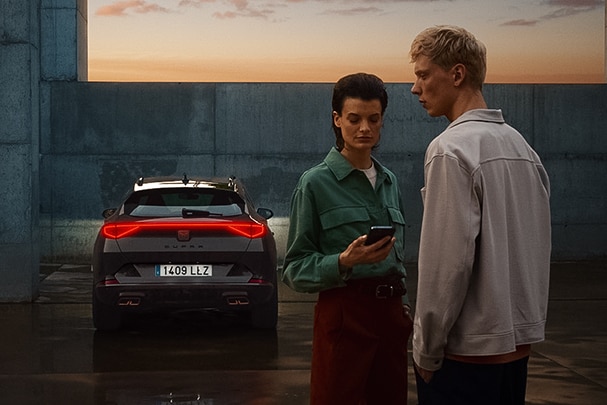 Un homme et une femme, qui tient un téléphone portable, se font face. En arrière-plan, une CUPRA est garée, ses phares sont allumés et on la voit de dos.