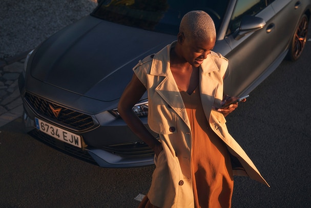 Une femme aux cheveux blonds très courts est appuyée contre une CUPRA bleue et regarde son téléphone portable.