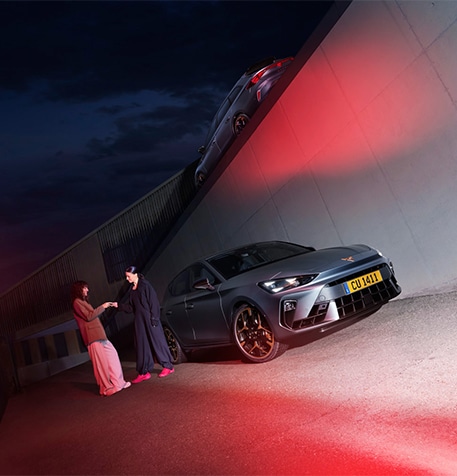 The 2024 Century bronze matte CUPRA Formentor parked on a reflective waterfront promenade. The sporty and aerodynamic new vehicle is positioned centre frame. The setting features futuristic architecture and Barcelona’s glowing city skyline at dusk.